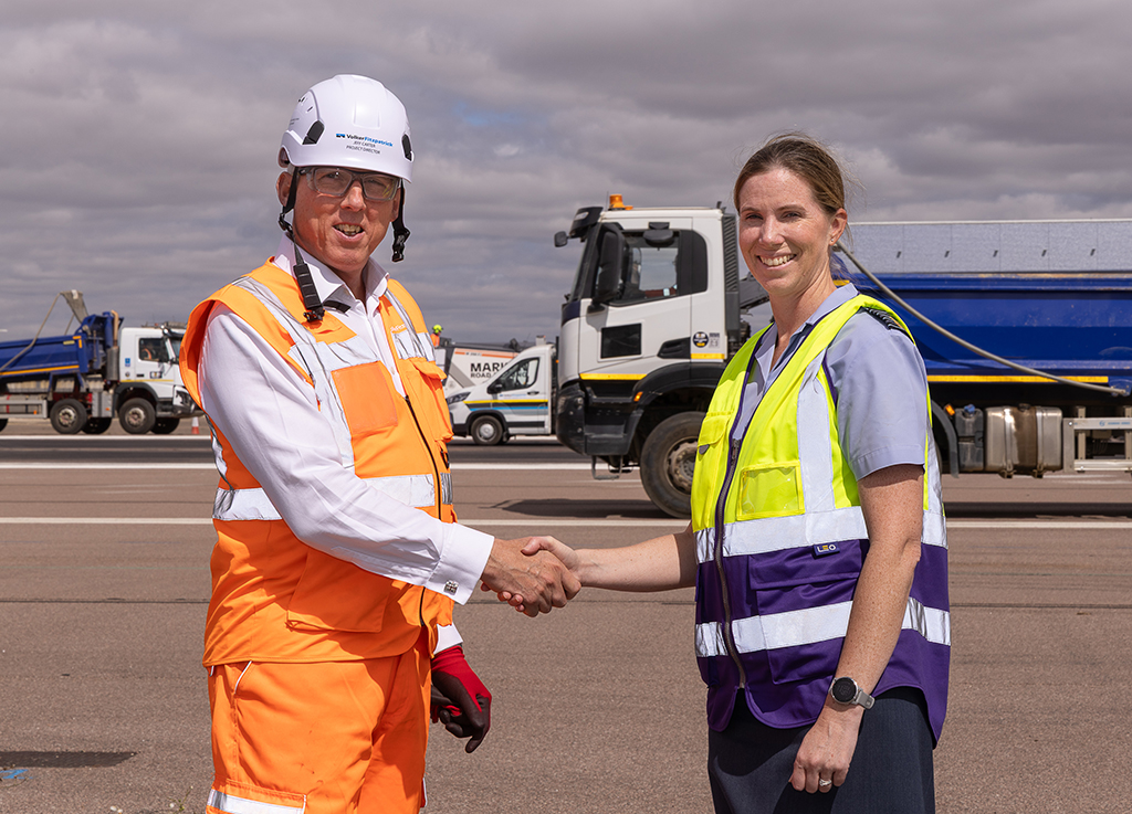 A runway works contractor wearing an orange high-visibility vest and a white hard hat, shakes hands with Group Captain Louise Henton who is wearing a purple and yellow high-visibility vest, as they stand doors on the runway asphalt surface.